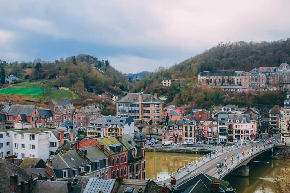 A picturesque aerial view of Dinant, Belgium, featuring a bridge adorned with colorful flags crossing the Meuse River. The town displays a charming mix of historic brick buildings and modern architecture, nestled between rolling green hills and forested slopes under a cloudy sky.