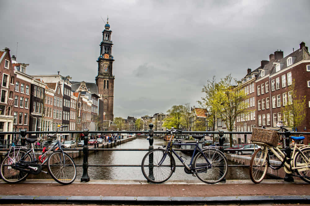 View of a canal in Amsterdam with bicycles parked along a bridge in the foreground. Traditional Dutch buildings line both sides of the water, and the Westerkerk church tower stands prominently in the background under a cloudy sky.