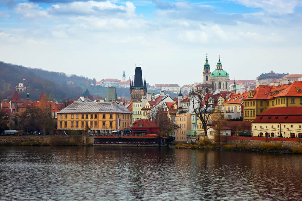 Scenic view of historic buildings along the Vltava River in Prague, Czech Republic, featuring red-tiled roofs, the Gothic tower of Charles Bridge, and the green dome and twin towers of St. Nicholas Church in the background under a partly cloudy sky.